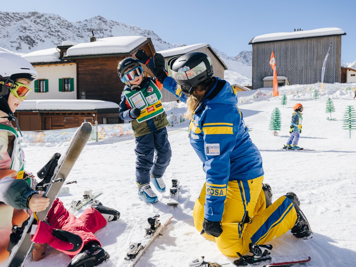Schneesportlehrerin der Skischule Arosa hilft Kindern im Kinderland beim Anziehen der Ski