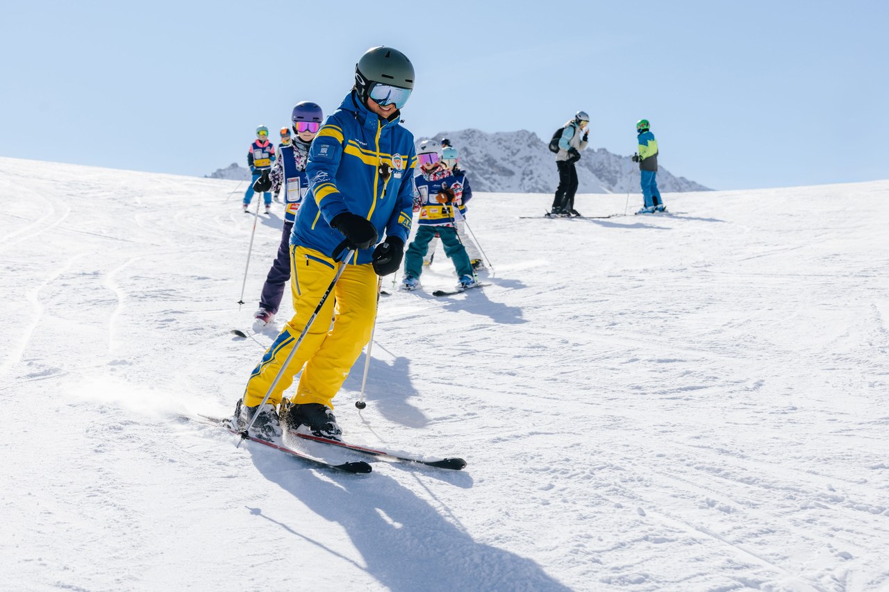 Kindergruppe im Blue-Star-Level fährt mit der Schneesportlehrerin über eine breite Piste in Arosa